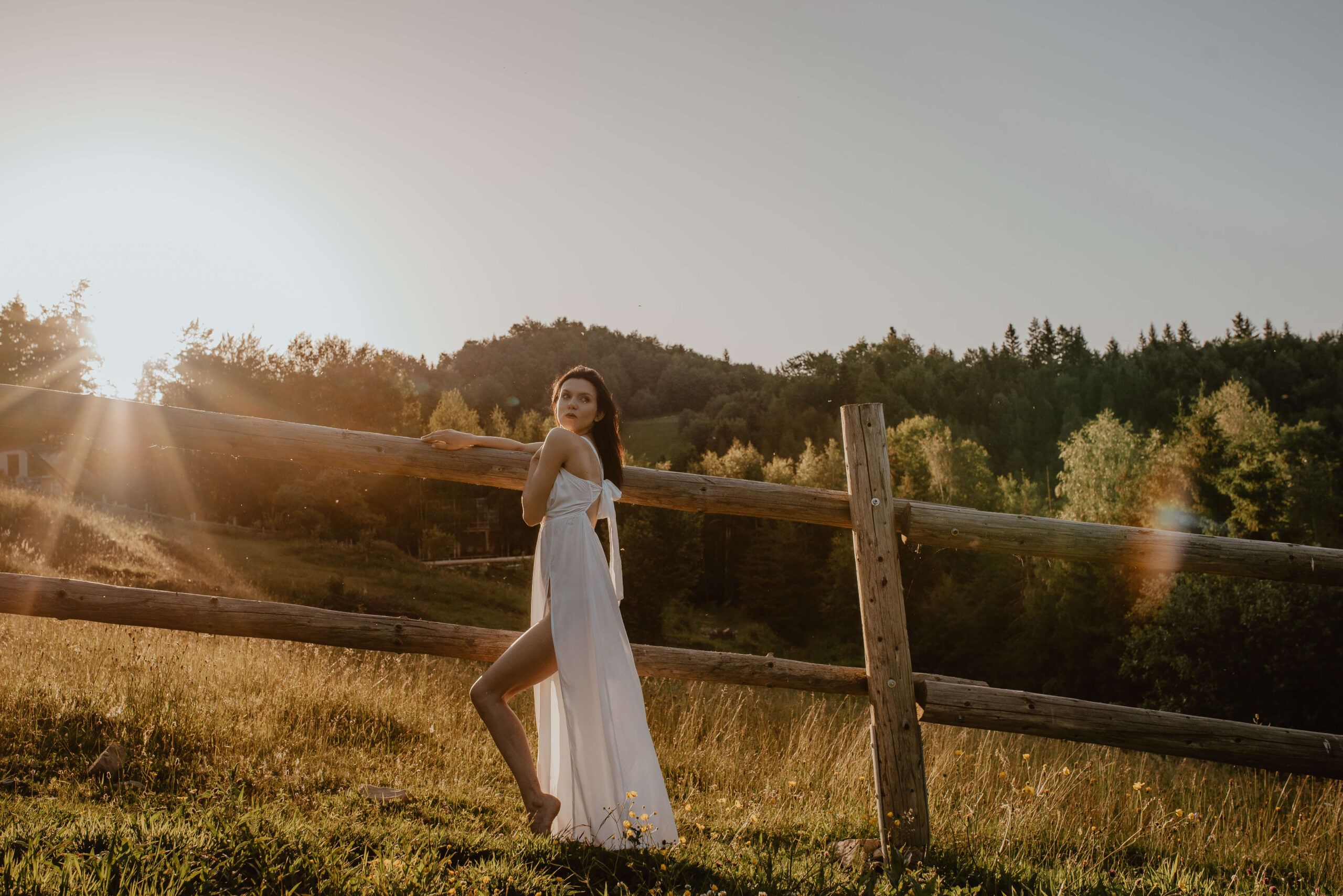 Eine natürliche und elegante Braut in einem leichten, fließenden Kleid, die den Sonnenaufgang in der Nähe eines Pferdestalls genießt und von der unberührten Schönheit der Natur umgeben ist.