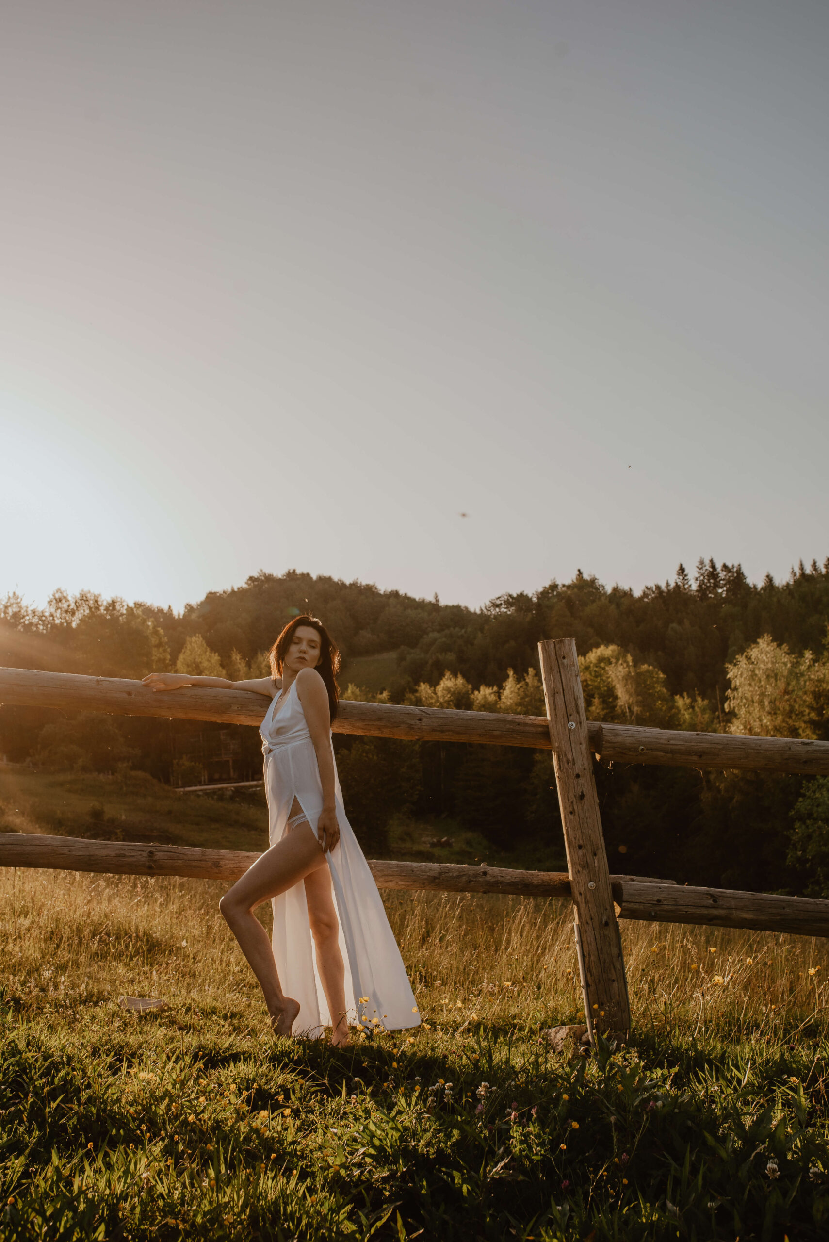 Die Braut in ihrem einfachen, luftigen Kleid steht vor einer hölzernen Pferdekoppel und genießt den Sonnenaufgang inmitten der unberührten Natur.
