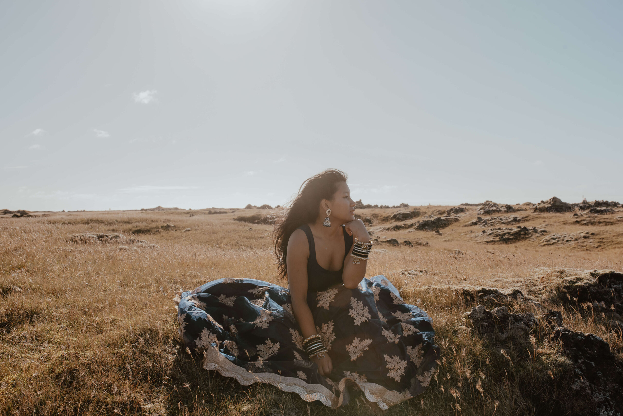 Das Foto zeigt eine junge Frau in einem traditionellen Kleid, die an einem windigen Sommertag auf einer isländischen Wiese steht und sich in der Sonne wärmt. Im Hintergrund des Fotos laufen Schafe in der Ferne umher. Die grünen Hügel und Berge, die den Hintergrund des Fotos bilden, erstrecken sich majestätisch gegen den blauen Himmel. Der Wind weht sanft durch das Gras und lässt es leicht hin- und herwogen. Die Farben des Fotos sind überwiegend Grün- und Blautöne, die die Schönheit und Unberührtheit der isländischen Landschaft widerspiegeln. Insgesamt strahlt das Bild eine gewisse Ruhe und Gelassenheit aus, die den Betrachter in einen Zustand der Entspannung versetzt. Die traditionelle Kleidung, die die junge Frau trägt, verleiht dem Foto eine gewisse kulturelle Bedeutung und verbindet die junge Frau mit ihrer Heimat und ihrer kulturellen Identität. Es ist offensichtlich, dass sie sich in diesem Moment in ihrer Umgebung wohl fühlt und in Einklang mit der Natur ist. Die Schafe im Hintergrund verleihen dem Foto eine weitere Dimension und veranschaulichen die enge Verbindung zwischen den Isländern und ihrer natürlichen Umgebung. Die junge Frau steht in einer selbstbewussten Haltung da, was ihre Stärke und Entschlossenheit zum Ausdruck bringt. Insgesamt ist das Foto ein beeindruckendes Porträt einer jungen Frau, die ihre Schönheit und ihre Verbindung zur Natur feiert.