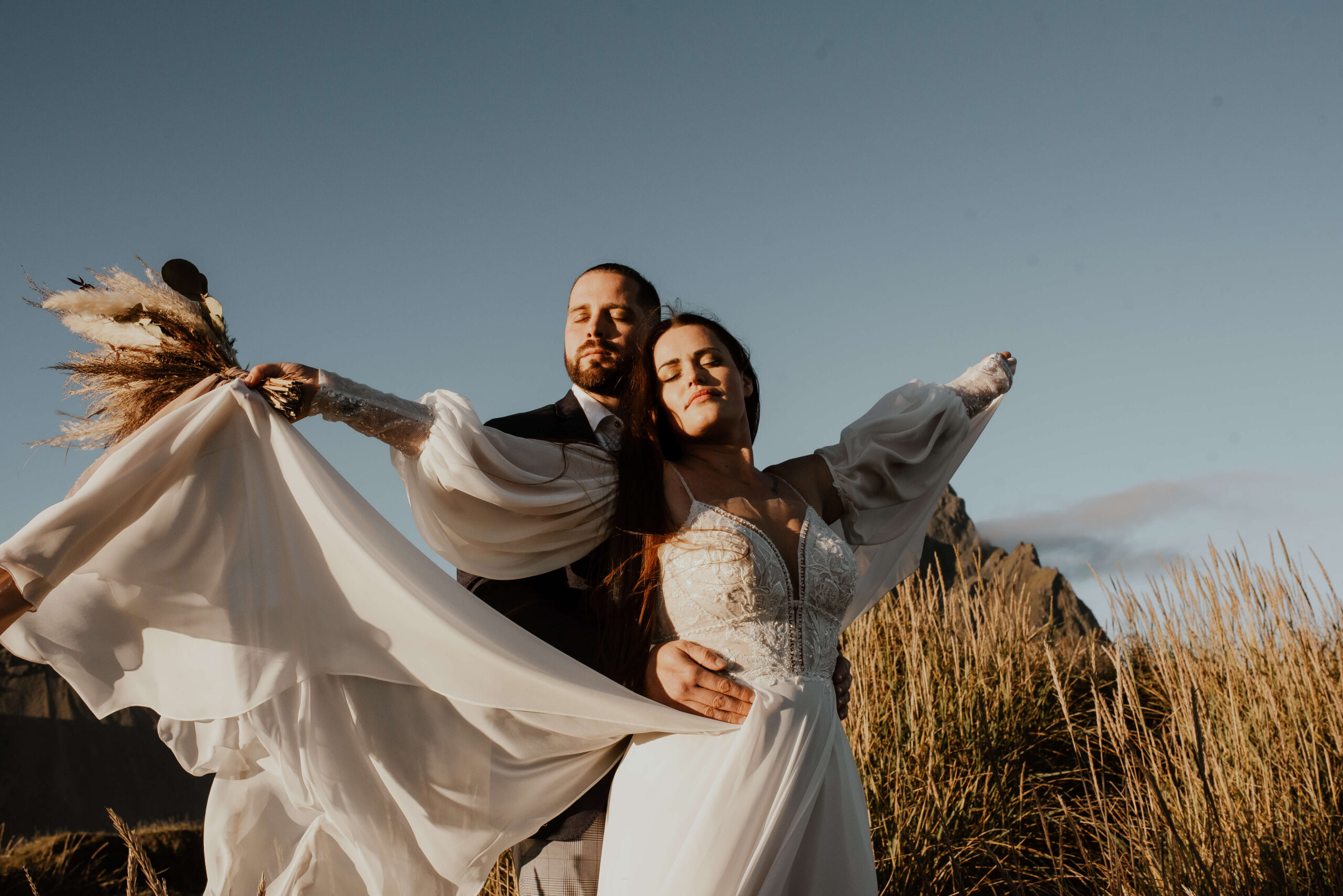 Auf dem Foto sieht man eine Hochzeitsreportage von Katarzyna Nord Fotografie. Das Bild zeigt ein junges Paar, das auf einem Gipfel in Island steht. Die Aussicht auf die umliegende Landschaft ist atemberaubend. Im Hintergrund sieht man eine Bergkette und den Himmel, der sich in verschiedenen Blautönen färbt. Das Paar steht Hand in Hand und schaut sich verliebt in die Augen. Die Braut trägt ein weißes Kleid und der Bräutigam einen dunklen Anzug. Beide haben einen glücklichen und zufriedenen Ausdruck auf dem Gesicht. Man kann spüren, dass sie diesen Moment auf dem Gipfel gemeinsam genießen und für immer in Erinnerung behalten werden.


