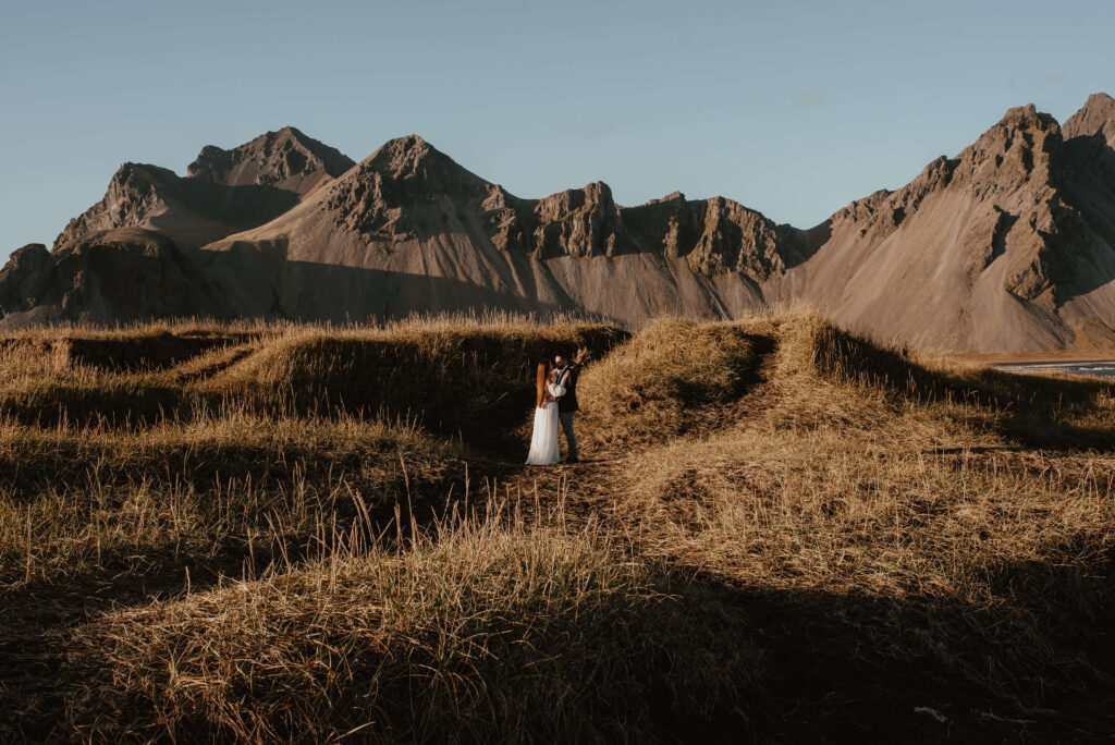 Auf dem Foto sieht man eine Hochzeitsreportage von Katarzyna Nord Fotografie. Das Bild zeigt ein junges Paar, das auf einem Gipfel in Island steht. Die Aussicht auf die umliegende Landschaft ist atemberaubend. Im Hintergrund sieht man eine Bergkette und den Himmel, der sich in verschiedenen Blautönen färbt. Das Paar steht Hand in Hand und schaut sich verliebt in die Augen. Die Braut trägt ein weißes Kleid und der Bräutigam einen dunklen Anzug. Beide haben einen glücklichen und zufriedenen Ausdruck auf dem Gesicht. Man kann spüren, dass sie diesen Moment auf dem Gipfel gemeinsam genießen und für immer in Erinnerung behalten werden.


