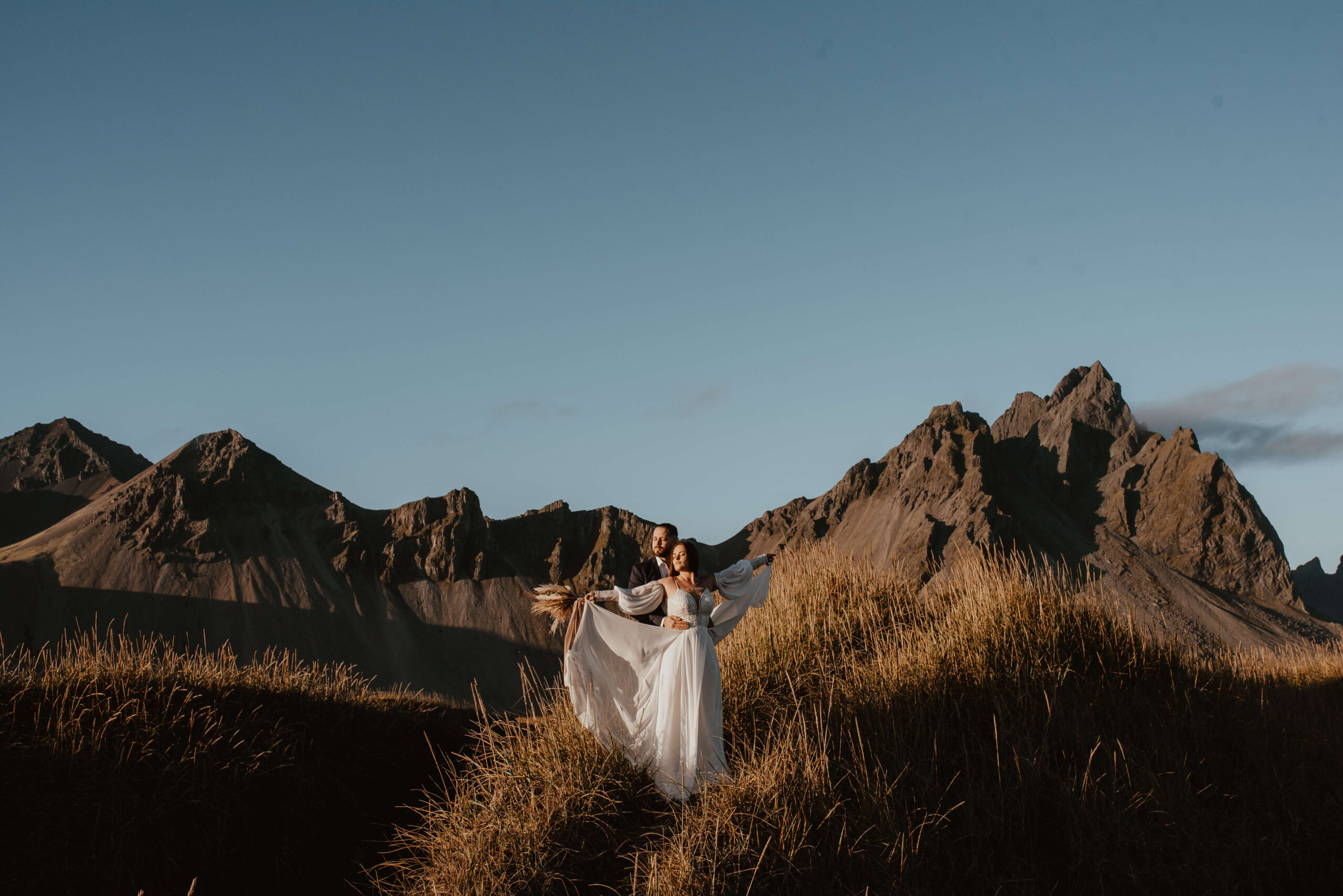 Auf diesem Foto sind ein junges Brautpaar in ihren Hochzeitskleidern auf Island zu sehen, an einem warmen Tag, mit im Hintergrund den majestätischen Bergen und dem Ozean. Das Paar umarmt sich lachend, während man das Rauschen der Wellen im Hintergrund hört. Diese Szene erinnert an eine legendäre Szene aus dem Film "Titanic". Das Foto zeigt die romantische und einzigartige Atmosphäre einer Hochzeit im Ausland. Katarzyna Nord Fotografie