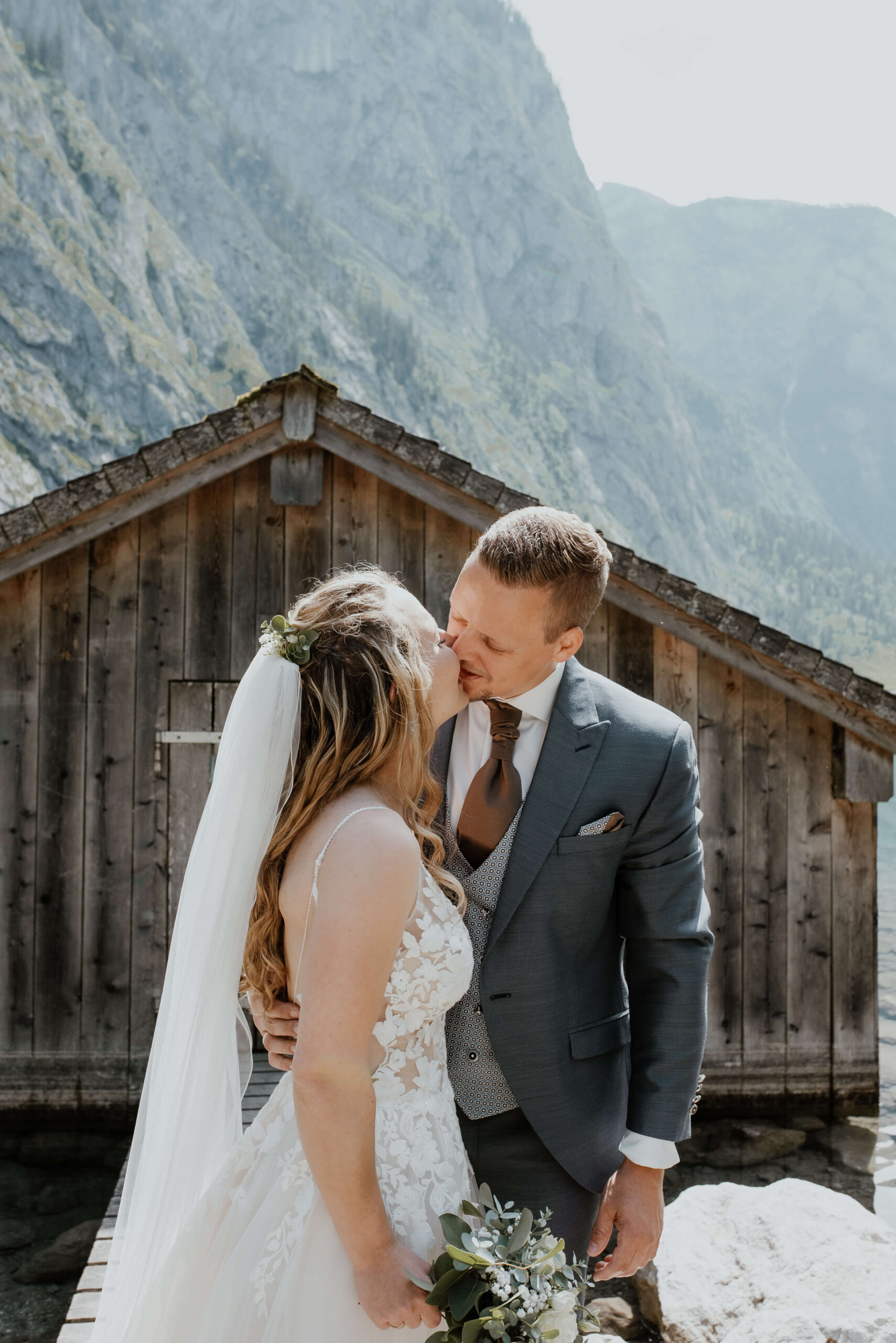Es ist eine Hochzeit am Konigsee, an einem heißen Tag im Mai. Die Braut trägt ihr Hochzeitskleid und küsst den Bräutigam. Beide stehen auf einer Holzbrücke und im Hintergrund sieht man eine Holzhütte und Berge. Der Hochzeitsfotograf Konigsee hält diesen besonderen Moment fest.